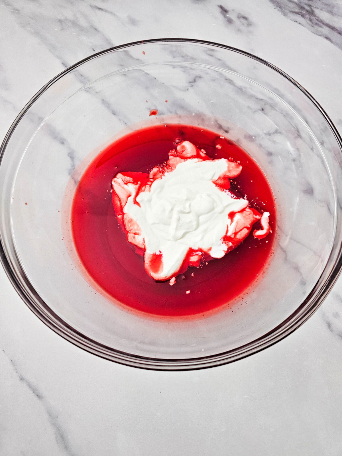 A scoop of smooth cottage cheese being added on top of the cooled strawberry gelatin in a glass bowl