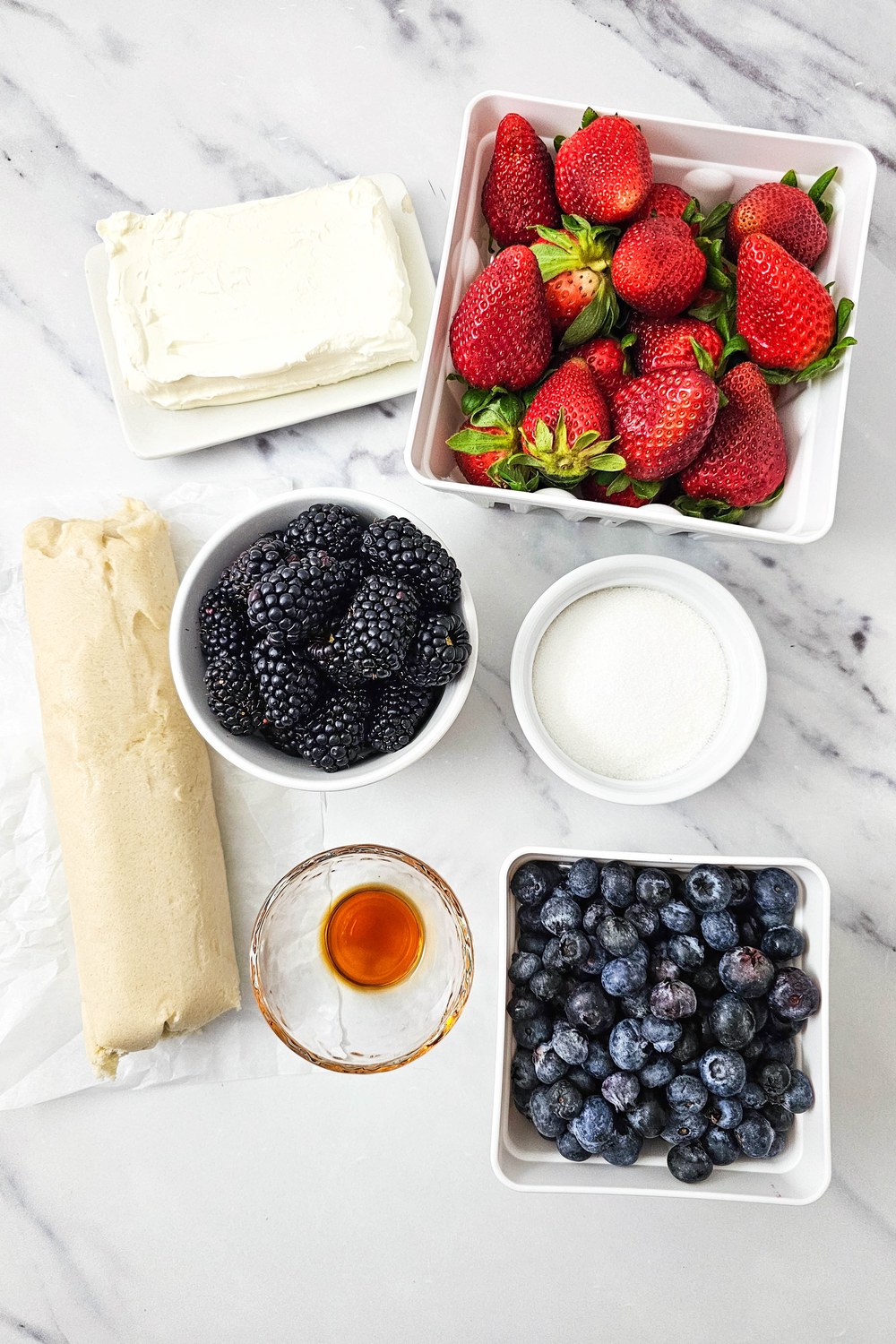 Flat lay of ingredients for red, white, and blue fruit pizza: cream cheese, sugar, vanilla, sugar cookie dough, strawberries, blueberries, and blackberries on a white marble surface